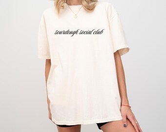 May include: A cream-coloured t-shirt featuring the text "sourdough social club" in a cursive font. The shirt has short sleeves and a relaxed fit. The model is wearing black shorts and a gold necklace.