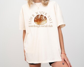 May include: Cream-coloured t-shirt with a watercolour illustration of a loaf of bread. The text "FLOUR. WATER. SALT." arches above the bread, with "sourdough social club" below. The shirt has short sleeves.