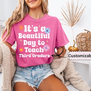 May include: Pink t-shirt with the text "It's a Beautiful Day to Teach Third Graders" in white with colorful flowers and stars. The text is customizable.