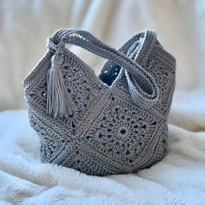 Könnte beinhalten: Handgefertigte, hellgraue Häkeltasche mit quadratischem Muster. Die Tasche hat einen Griff und eine dekorative Quaste. Die Tasche steht auf einer weißen, flauschigen Oberfläche.