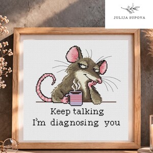 Puede incluir: Un patrón de punto de cruz en un marco de madera que muestra una rata gris con orejas rosas sosteniendo una taza morada con vapor. El texto debajo de la rata dice, Keep talking I'm diagnosing you. El diseño está sobre un fondo de tela blanca.