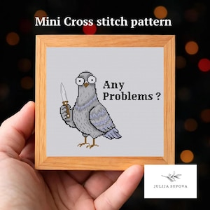 May include: A square wooden frame containing a cross stitch pattern of a grey pigeon with wide eyes holding a small knife. The text Any Problems? is stitched in black next to the bird. The background of the stitched area is light grey.