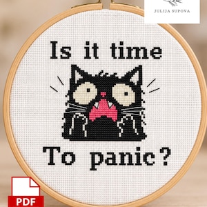 May include: A cross stitch pattern in a wooden embroidery hoop featuring a black cat with wide eyes and a pink mouth on white fabric. The text reads Is it time To panic? The design includes a PDF label and the brand name Julija Supova.