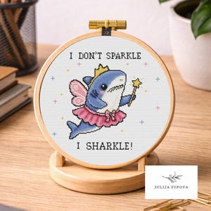 Può includere: Un telaio da ricamo a punto croce con uno squalo dei cartoni animati vestito da fata, con una corona e un tutù, che tiene in mano una bacchetta a stella. Il testo recita "I DON'T SPARKLE I SHARKLE!" Il telaio è esposto su un supporto di legno.