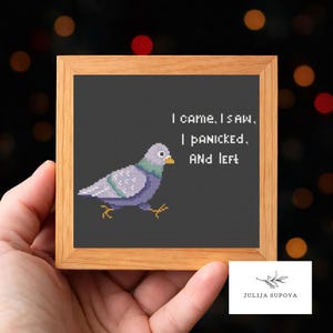 May include: A framed cross-stitch featuring a pixelated pigeon with the text "I came, I saw, I panicked, AND left." The bird is primarily gray and purple, with yellow feet and a yellow beak. The frame is made of light-colored wood.
