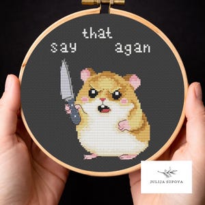 Puede incluir: Bordado de punto de cruz en un aro de madera con un hámster enfadado sosteniendo un cuchillo. El texto "say that again" está bordado encima del hámster. El hámster es marrón, beige y rosa, con un fondo negro.