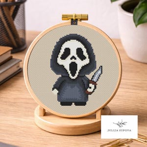 Può includere: Un ricamo a punto croce in un telaio di legno raffigura il personaggio dei cartoni animati Ghostface dei film Scream, con un coltello. Il personaggio indossa una veste nera con una maschera bianca. Il ricamo è esposto su un supporto di legno.