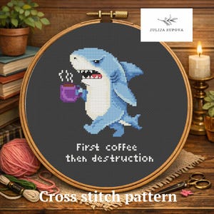May include: Cross-stitch pattern showing a blue cartoon shark with a purple coffee mug, and the text "First coffee then destruction". The words "Cross stitch pattern" are at the bottom. The design is set in a wooden embroidery hoop.