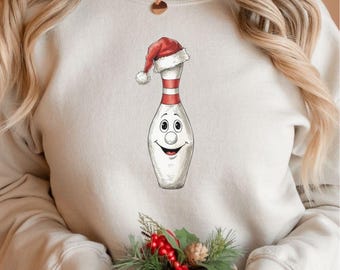 May include: A cream-coloured sweatshirt with a cartoon bowling pin wearing a Santa hat. The bowling pin has a smiling face and red and white stripes. The sweatshirt is a festive holiday design.