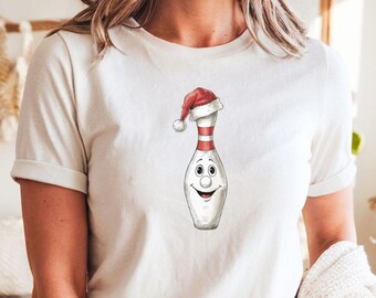 May include: White t-shirt featuring a cartoon bowling pin wearing a Santa hat. The pin has a smiling face and red and white stripes. The design is a festive, holiday-themed graphic.