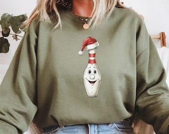 May include: An olive green sweatshirt with a cartoon bowling pin wearing a Santa hat. The bowling pin has a smiling face and a red and white striped design. The sweatshirt is a casual, long-sleeved top, perfect for the festive season.