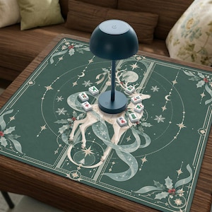 Può includere: Un tappetino quadrato verde scuro con un'illustrazione di un cervo bianco, foglie di agrifoglio e stelle, sormontato da una lampada blu scuro a forma di fungo e diverse piccole tessere bianche del mahjong con caratteri rossi e verdi.