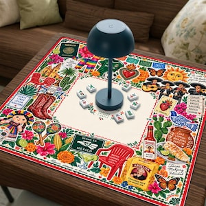 May include: A square game mat featuring a colorful Mexican-themed illustration with icons like maracas, cowboy boots, cacti, a bottle of hot sauce, and a lamp in the center. The mat includes scattered mahjong tiles and text such as Mexico, Abuelita, and Buena Compañía Juega Juega.