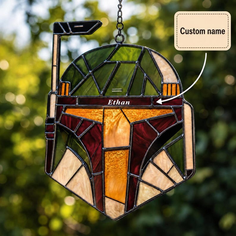 May include: A stained glass suncatcher shaped like a helmet with a rangefinder, featuring green, yellow, maroon, and cream glass panels. The horizontal band across the center includes the custom name Ethan in white text, suspended by a metal chain.