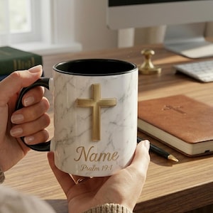 May include: A white marble-effect mug featuring a gold cross. The mug has a black interior and handle. The text "Name" and "Psalm 19:1" are printed in gold script on the mug.
