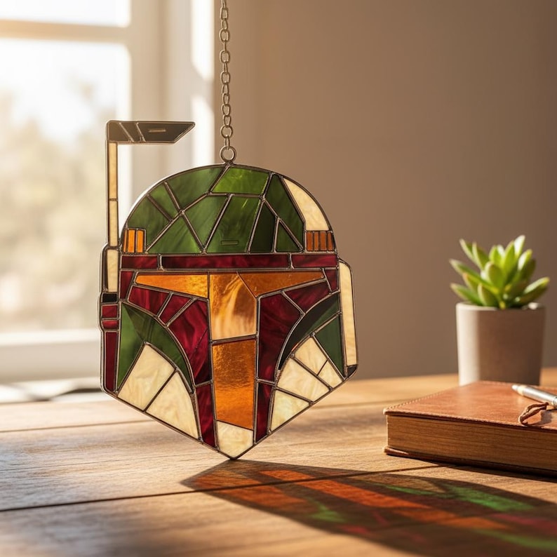 May include: A stained glass suncatcher shaped like a helmet with a T-shaped visor. The piece features geometric panels in shades of green, deep red, and amber, held together by dark metal solder lines. A metal chain is attached to the top for hanging.