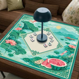 Könnte beinhalten: Eine quadratische Mahjong-Matte mit einer grünen Lotus-Teich-Illustration, rosa Blüten, einem Boot und einem Stück Wassermelone. Eine dunkelblaue pilzförmige Lampe steht mittig auf einem cremefarbenen Quadrat, umgeben von acht verstreuten Mahjong-Steinen.