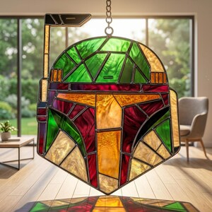 May include: A stained glass suncatcher shaped like a helmet with a rangefinder. The piece features geometric panels in shades of green, deep red, and amber, held together by dark metal solder lines. It hangs from a small metal chain at the top.