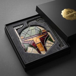 May include: A stained glass helmet ornament featuring a geometric design in shades of green, maroon, orange, and cream, with a dark metal chain attached. The piece is presented in a black gift box with a gold sticker reading Handmade.