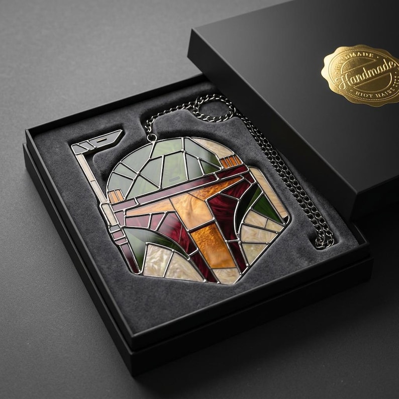 May include: A stained glass helmet ornament featuring a geometric design in shades of green, maroon, orange, and cream, with a dark metal chain attached. The piece is presented in a black gift box with a gold sticker reading Handmade.