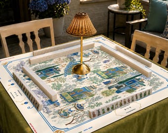 Puede incluir: Un tapete de mahjong cuadrado con superficie blanca, decorado con ilustraciones azules y verdes de edificios y árboles tradicionales, rodeado por cuatro filas de fichas blancas. Una pequeña lámpara dorada con pantalla tejida se sitúa en el centro.