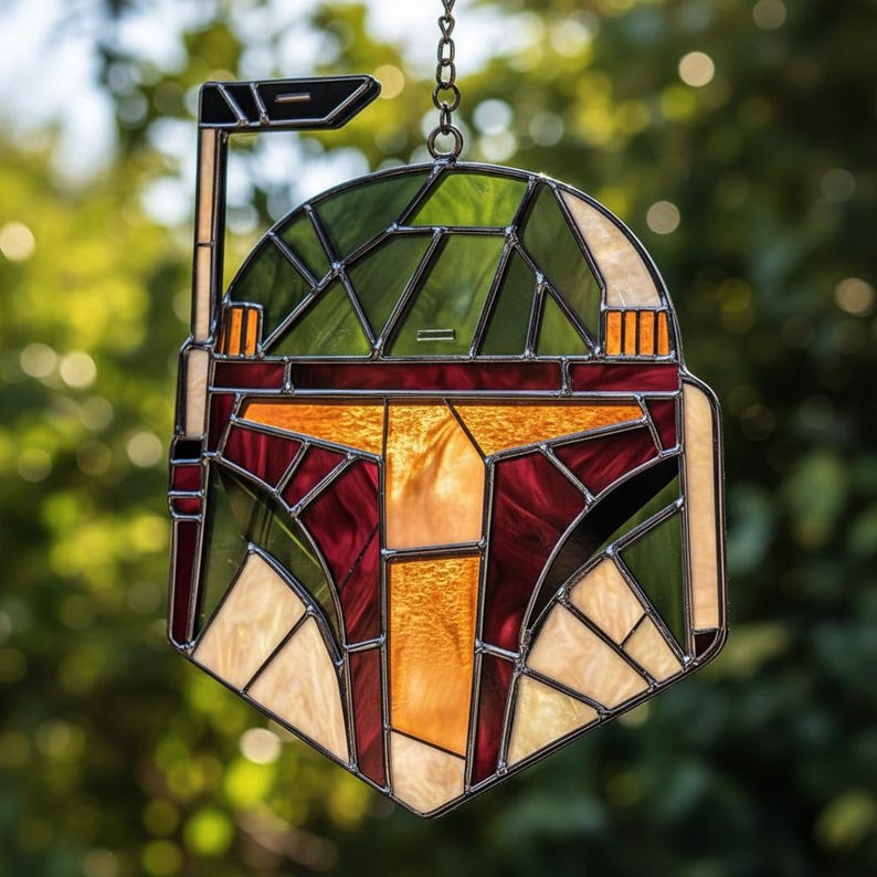May include: A stained glass suncatcher shaped like a helmet with a T-shaped visor. The piece features dark green, deep red, and cream-colored glass panels held together by dark metal solder lines, including an antenna on the left side.