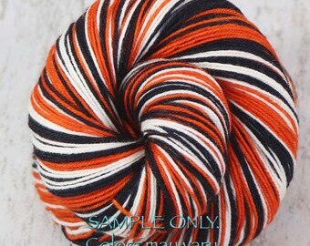 May include: A skein of yarn with a striped pattern in orange, black, and white. The yarn is tightly wound, showing the texture of the fibers. Text on the image reads "SAMPLE ONLY. Colors may vary due to fiber content."