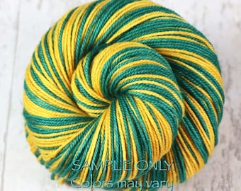 Könnte beinhalten: Ein rundes Garnknäuel mit einem lebendigen, abwechselnd gestreiften Muster aus tiefem Waldgrün und hellem Goldgelb. Der Text "SAMPLE ONLY. Colors may vary due to fiber content." ist in einer dunkelblauen Schrift mittig aufgedruckt.