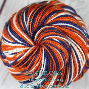 May include: A ball of yarn in shades of orange, navy blue, and white. The yarn is tightly wound, with the colors arranged in a striped pattern. The text "SAMPLE ONLY. Colors may vary due to fiber content" is visible.