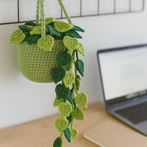 Könnte beinhalten: Ein handgefertigter hängender Häkel-Pflanztopf in Grüntönen. Der Topf ist mit gehäkelten Blättern in verschiedenen Grüntönen gefüllt, wobei einige Blätter nach unten fallen. Der Topf wird an einem schwarzen Haken aufgehängt.