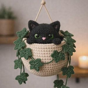 May include: A crocheted hanging basket with a black cat inside. The cat has green eyes and a pink nose. The basket is beige, with green ivy leaves. It hangs from a beige rope. The cat is the focal point.