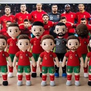 May include: A group of crocheted dolls representing the Portugal national soccer team, wearing red jerseys with green shorts. The dolls have various skin tones and hairstyles, with some sporting beards. A framed photo of the team is in the background.