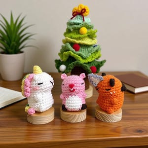 May include: A collection of handmade crochet figures, including a white unicorn with a rainbow mane, a pink hamster, an orange fox, and a green Christmas tree. Each figure is mounted on a small wooden base, creating a whimsical display.