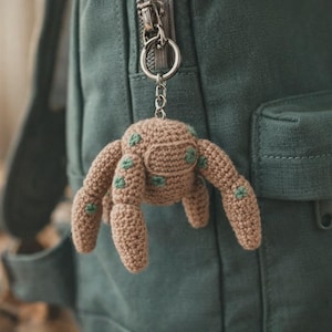 Könnte beinhalten: Ein gehäkelter Spinnen-Schlüsselanhänger in Beige mit grünen Akzenten, der an einem silbernen Schlüsselring an einem grünen Rucksack hängt. Die Spinne hat acht Beine und einen detaillierten Körper, der Handarbeit zeigt.