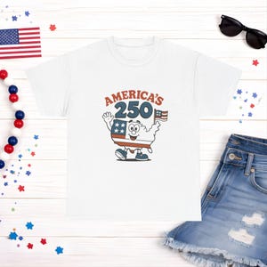 May include: White t-shirt featuring a cartoon map of the United States with a waving hand and holding a flag. The text "America's 250" is above the cartoon. Sunglasses and denim shorts are in the background.