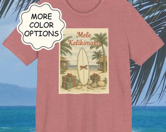 May include: A short-sleeved, dusty rose t-shirt featuring a vintage-style graphic. The graphic depicts a surfboard with a bow, gifts, and palm trees, with the words "Mele Kalikimaka". The shirt also has a "More Color Options" button.