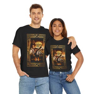 Peut inclure: T-shirts noirs avec un motif d'affiche de film dans un cadre doré. L'affiche représente des personnages avec des fusils sur fond de désert. Le mot "MUMMY" est visible en bas du motif.
