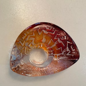 May include: A clear glass ring holder with a brown, orange, and white swirl pattern. The holder is shaped like a teardrop and has a smooth, rounded edge.