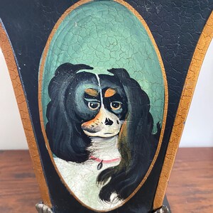 K&ouml;nnte beinhalten: Eine schwarz-goldene dekorative Vase mit einem gemalten ovalen Portr&auml;t eines Hundes. Der Hund hat schwarzes Fell, ein wei&szlig;es Gesicht und ein rosa Halsband. Die Vase hat eine antike Oberfl&auml;che und verzierte Metallf&uuml;&szlig;e.