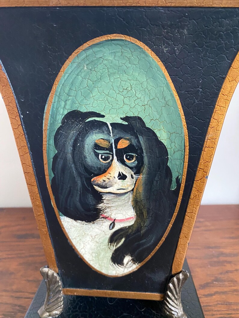 K&ouml;nnte beinhalten: Eine schwarz-goldene dekorative Vase mit einem gemalten ovalen Portr&auml;t eines Hundes. Der Hund hat schwarzes Fell, ein wei&szlig;es Gesicht und ein rosa Halsband. Die Vase hat eine antike Oberfl&auml;che und verzierte Metallf&uuml;&szlig;e.