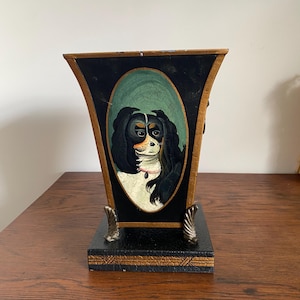 K&ouml;nnte beinhalten: Eine dekorative schwarze und goldene Vase mit einem gemalten Portr&auml;t eines Cavalier King Charles Spaniels. Der Hund hat schwarzes, wei&szlig;es und braunes Fell, vor einem gr&uuml;nen ovalen Hintergrund. Die Vase hat einen quadratischen Sockel und verzierte goldene Akzente.