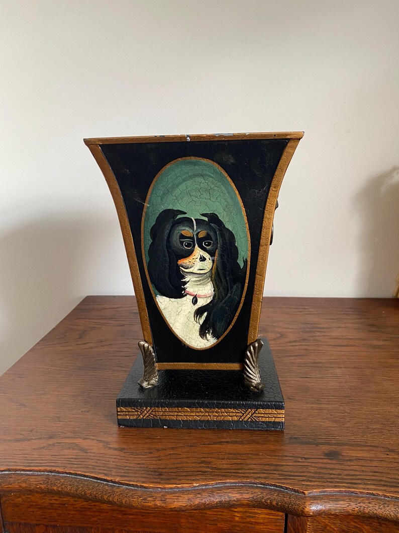 K&ouml;nnte beinhalten: Eine dekorative schwarze und goldene Vase mit einem gemalten Portr&auml;t eines Cavalier King Charles Spaniels. Der Hund hat schwarzes, wei&szlig;es und braunes Fell, vor einem gr&uuml;nen ovalen Hintergrund. Die Vase hat einen quadratischen Sockel und verzierte goldene Akzente.