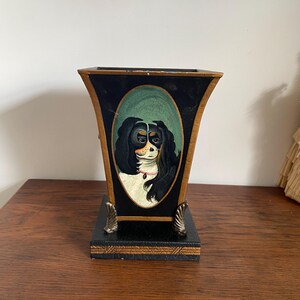 K&ouml;nnte beinhalten: Eine schwarz-goldene dekorative Vase mit einem gemalten Portr&auml;t eines Cavalier King Charles Spaniel. Die Vase hat einen rechteckigen Sockel und eine ausgestellte Oberseite mit goldenen Akzenten und dekorativen F&uuml;&szlig;en.