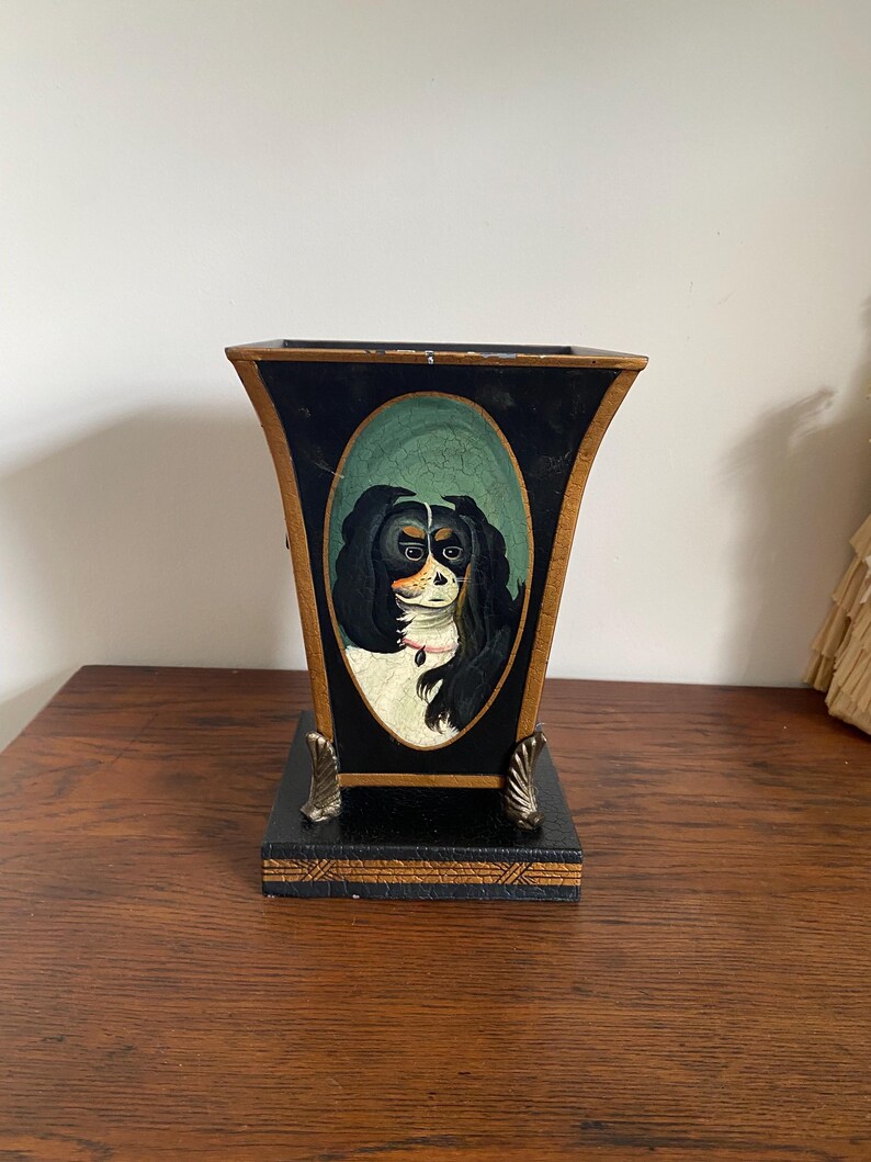 K&ouml;nnte beinhalten: Eine schwarz-goldene dekorative Vase mit einem gemalten Portr&auml;t eines Cavalier King Charles Spaniel. Die Vase hat einen rechteckigen Sockel und eine ausgestellte Oberseite mit goldenen Akzenten und dekorativen F&uuml;&szlig;en.