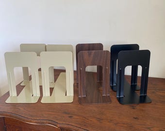 May include: A collection of metal bookends in various colors, including beige, dark wood grain, and black. The bookends have a simple, modern design with a rectangular shape and a support base. They are arranged on a wooden surface.