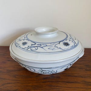 May include: A white ceramic lidded bowl with blue floral and swirl designs. The bowl has a rounded shape with a small handle on the lid. The rim of the bowl and lid are accented with a thin gold band. The bowl is sitting on a wooden surface.