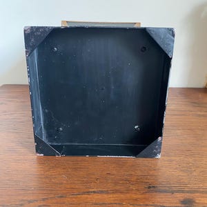 K&ouml;nnte beinhalten: Eine schwarze Metallbox mit rechteckiger Form. Die Box weist eine leicht abgenutzte Optik mit sichtbaren Abnutzungserscheinungen an den Ecken auf. Der Innenraum ist ebenfalls schwarz, und die Box steht auf einer Holzoberfl&auml;che.