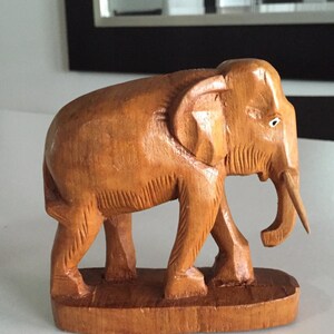May include: A hand-carved wooden elephant figurine with a light brown finish. The elephant is standing on a wooden base and has a long, thin trunk.