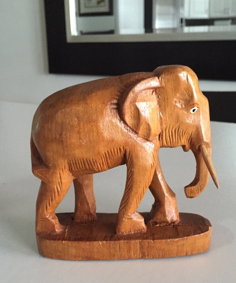 May include: A hand-carved wooden elephant figurine with a light brown finish. The elephant is standing on a wooden base and has a long, thin trunk.
