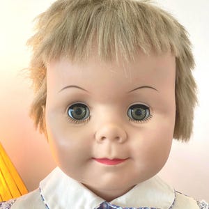 May include: A vintage doll with blonde hair, blue eyes, and a red mouth. She is wearing a white collared shirt with a blue and red plaid pattern.